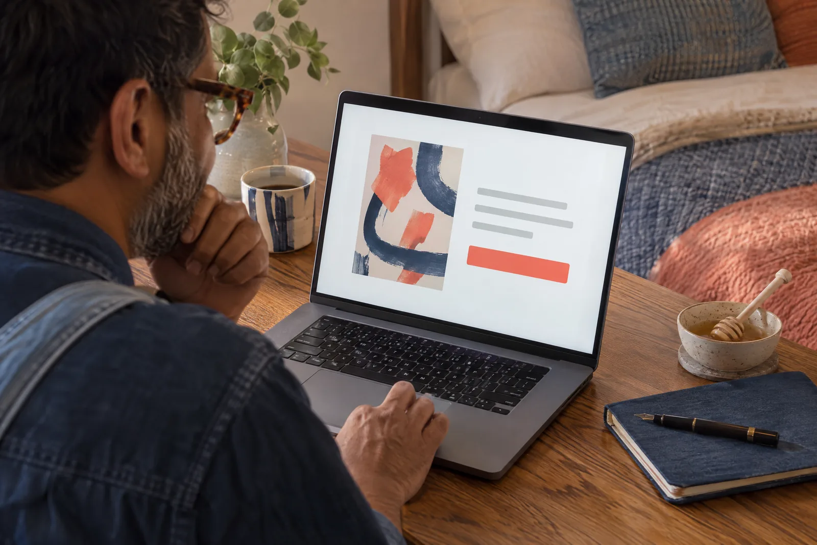 A south asian man in his forties with tortoiseshell glasses in a denim overalls at a bedroom alcove looks at a screen showing a payment product for sale, with a honey dipper beside them.