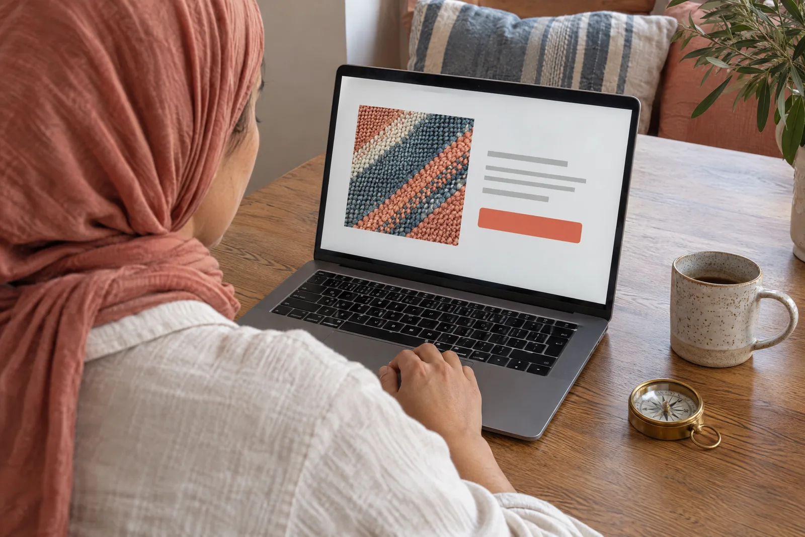 A woman in her thirties wearing a headscarf in a cream linen shirt at a dining alcove looks at a screen showing a general product for sale, with a brass compass beside them.