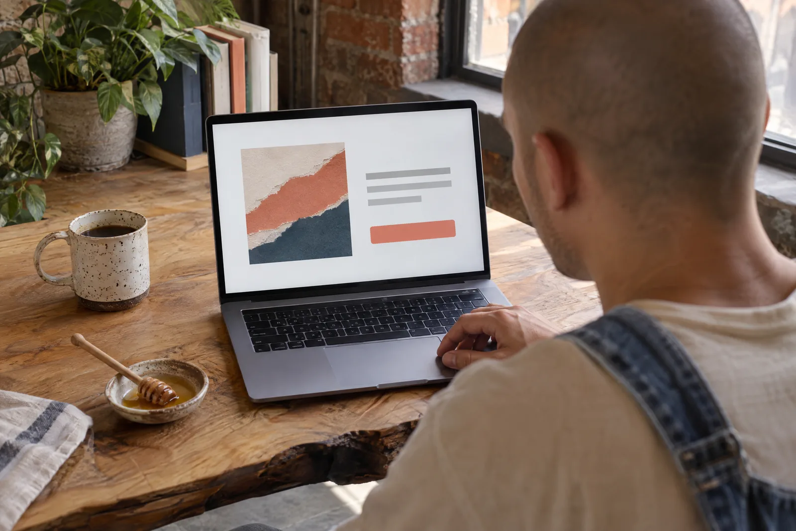 A man in his twenties with a shaved head in a denim overalls at a loft corner looks at a screen showing a general product for sale, with a honey dipper beside them.