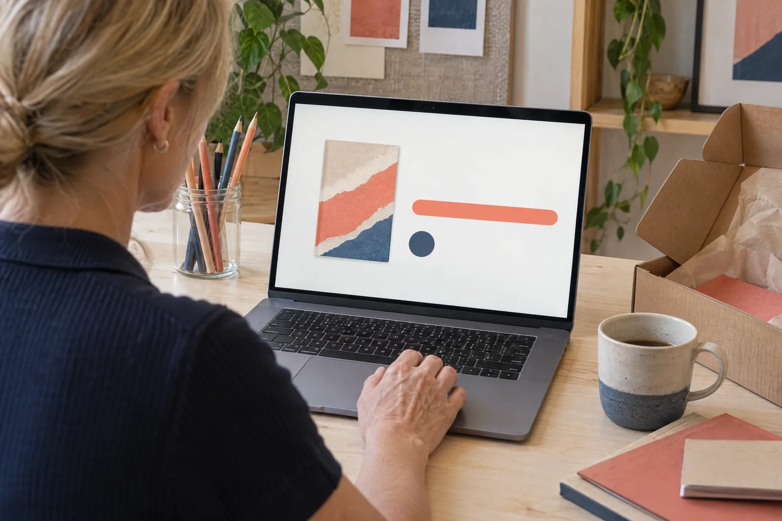 A woman in her forties with blonde hair tied back in a navy knit polo at a editing bay looks at a screen showing a delivery product for sale, with a glass jar of pencils beside them.