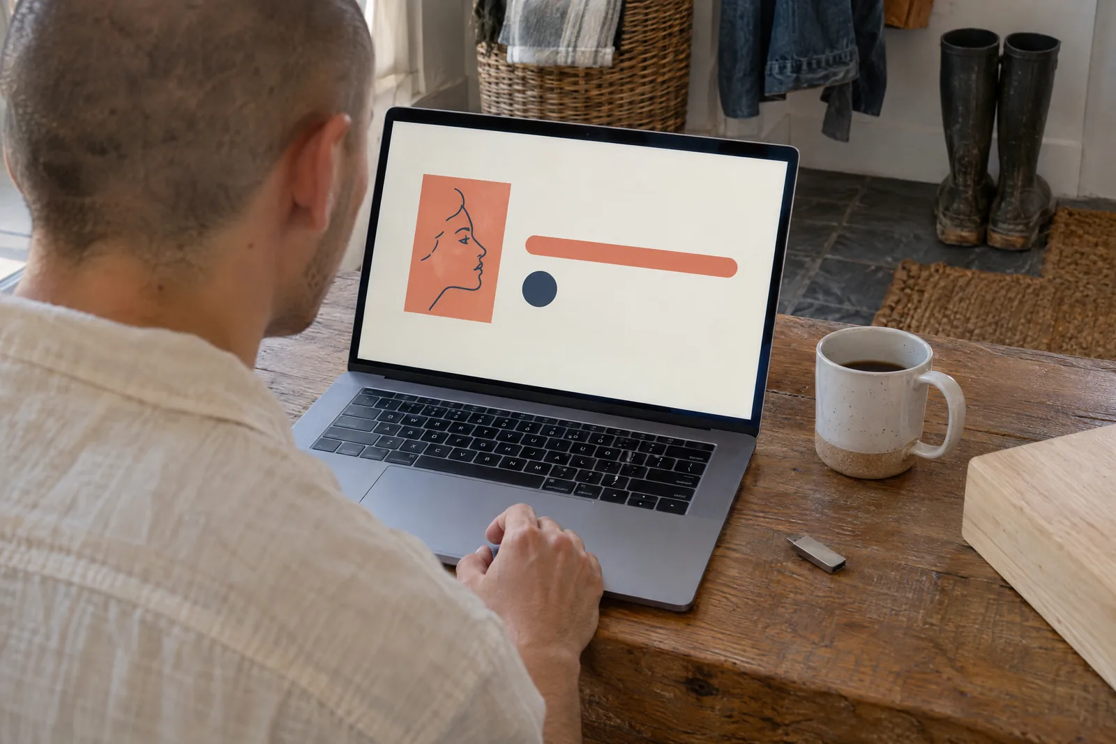 A man in his twenties with a shaved head in a cream linen shirt at a mudroom bench looks at a screen showing a software product for sale, with a pale wooden block beside them.