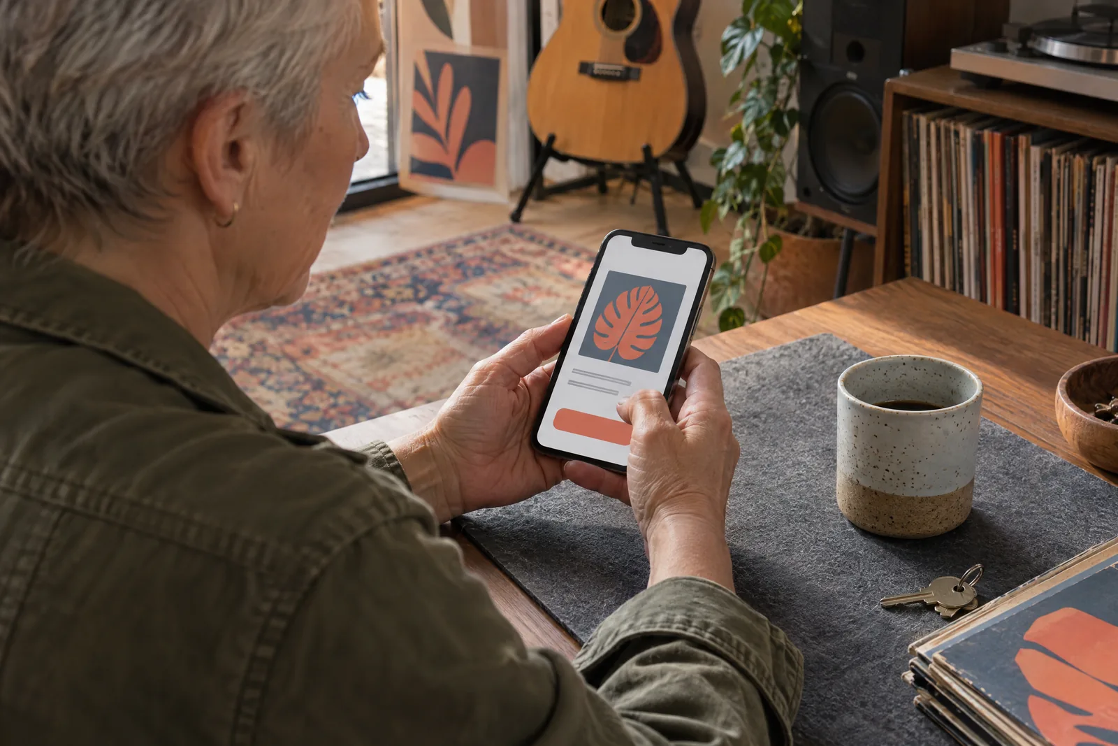 An older woman with cropped gray hair in a olive field jacket at a music room looks at a screen showing a general product for sale, with a house key beside them.