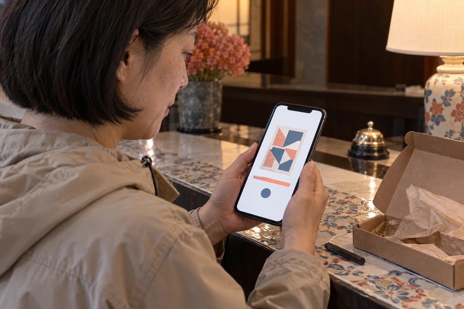 An east asian woman in her forties with a short bob in a sand rain jacket at a hotel desk looks at a screen showing a delivery product for sale, with a charcoal stick beside them.