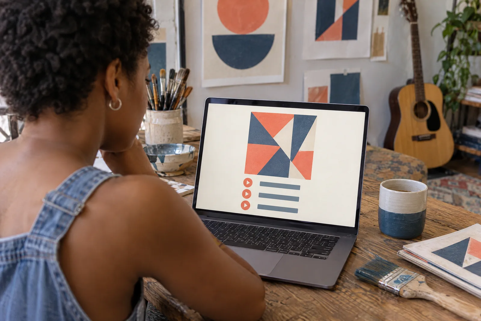 A black woman in her twenties with short natural hair in a denim overalls at a print studio looks at a screen showing a music product for sale, with a wide paintbrush beside them.