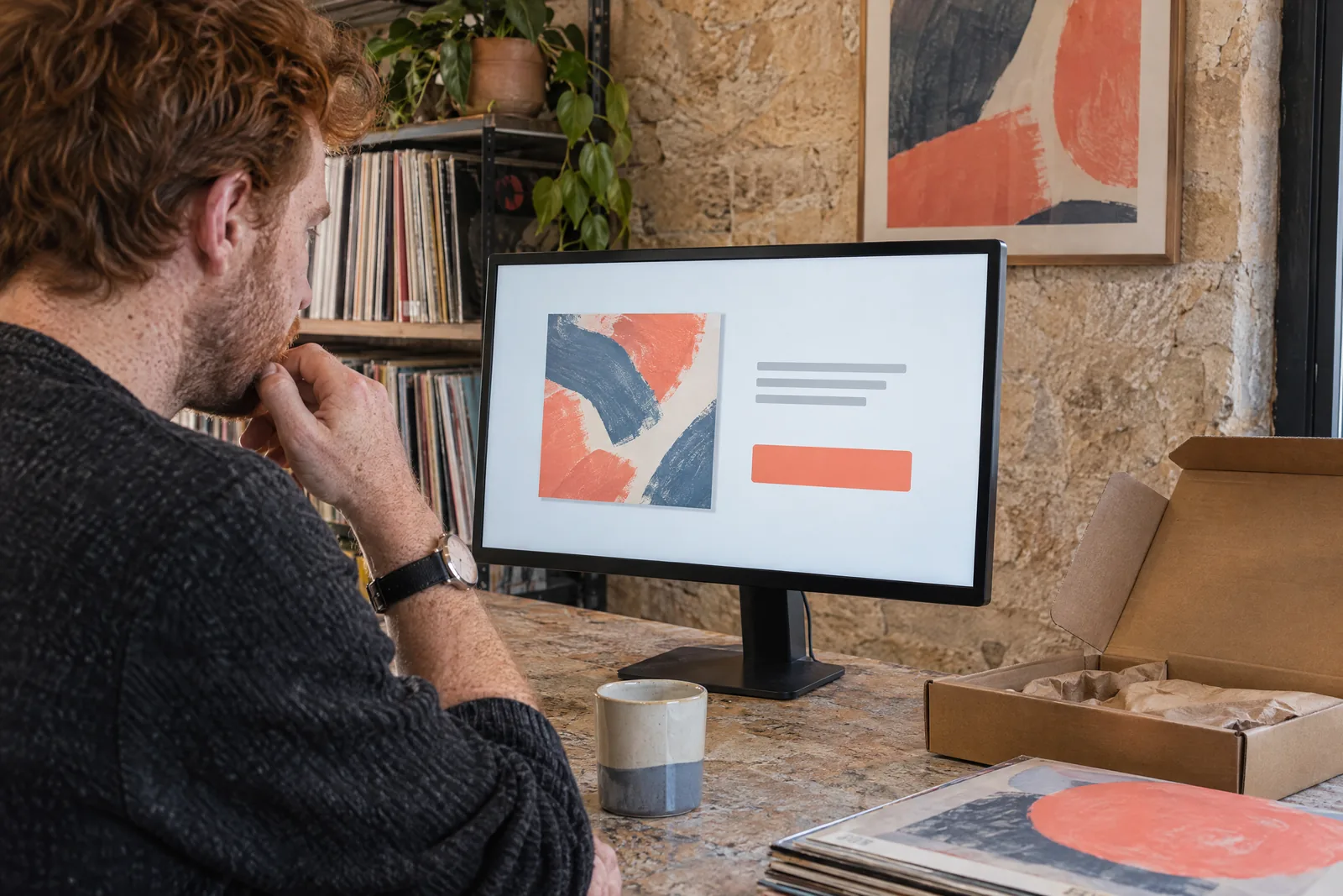 A man in his thirties with red hair and freckles in a charcoal sweater at a record nook looks at a screen showing a delivery product for sale, with a wristwatch with a blank face beside them.