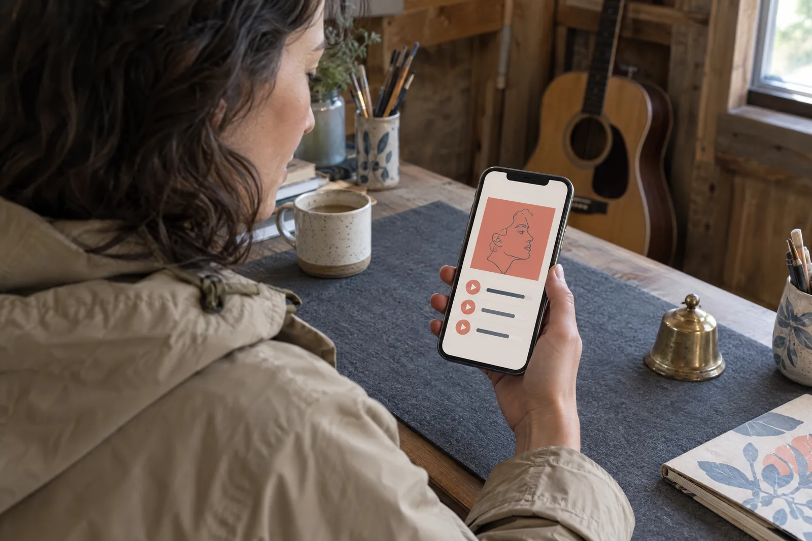 A woman in her thirties with dark wavy hair in a sand rain jacket at a craft barn looks at a screen showing a music product for sale, with a small brass bell beside them.