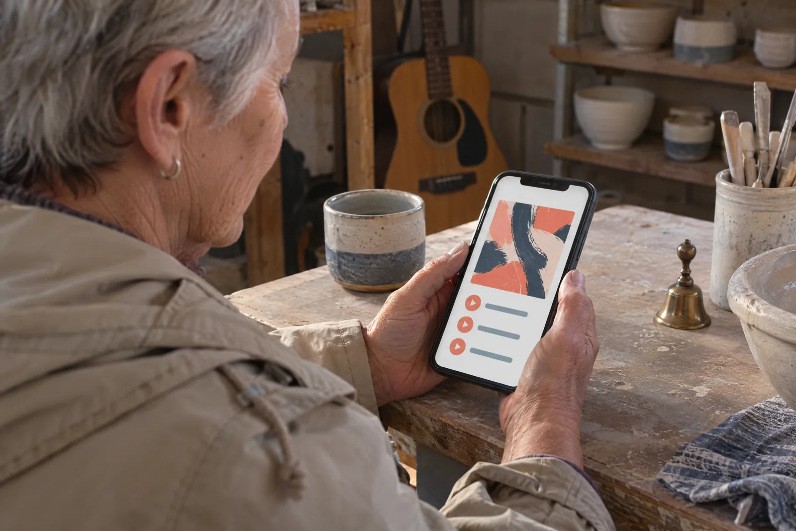 An older woman with cropped gray hair in a sand rain jacket at a pottery shed looks at a screen showing a music product for sale, with a small brass bell beside them.