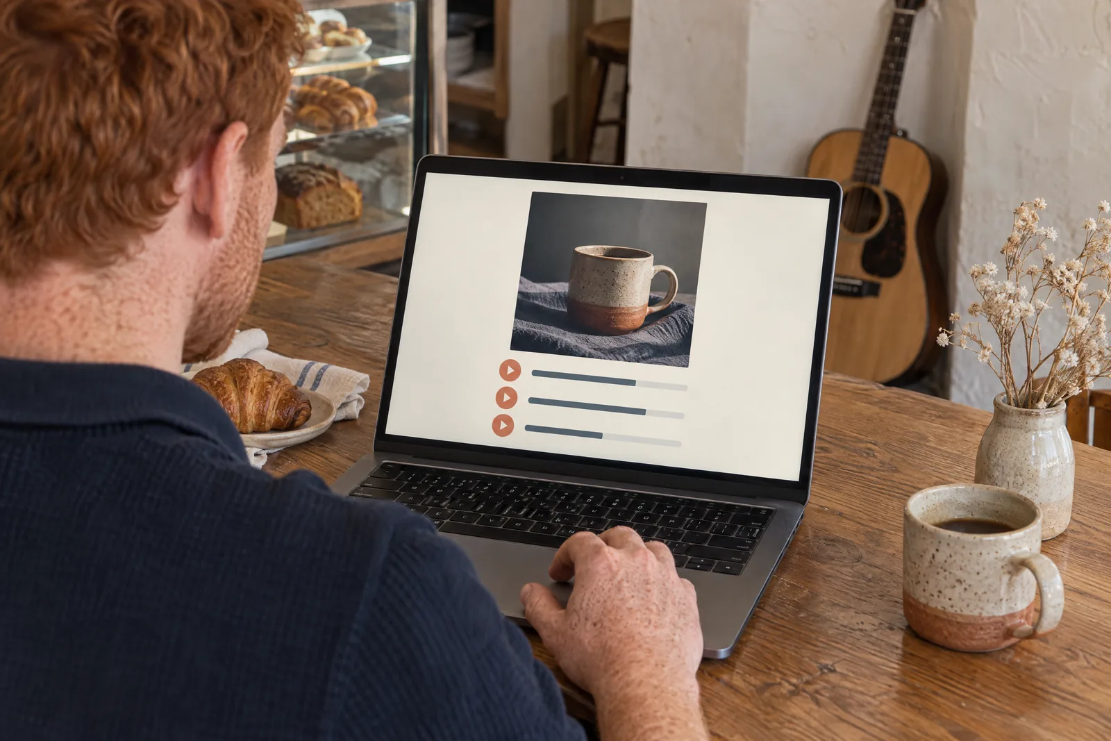 A man in his thirties with red hair and freckles in a navy knit polo at a bakery side counter looks at a screen showing a music product for sale, with a dried flower beside them.