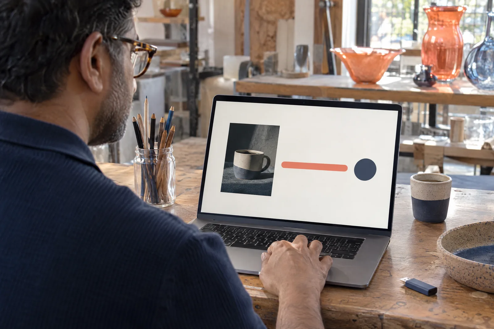 A south asian man in his forties with tortoiseshell glasses in a navy knit polo at a glass studio looks at a screen showing a software product for sale, with a glass jar of pencils beside them.