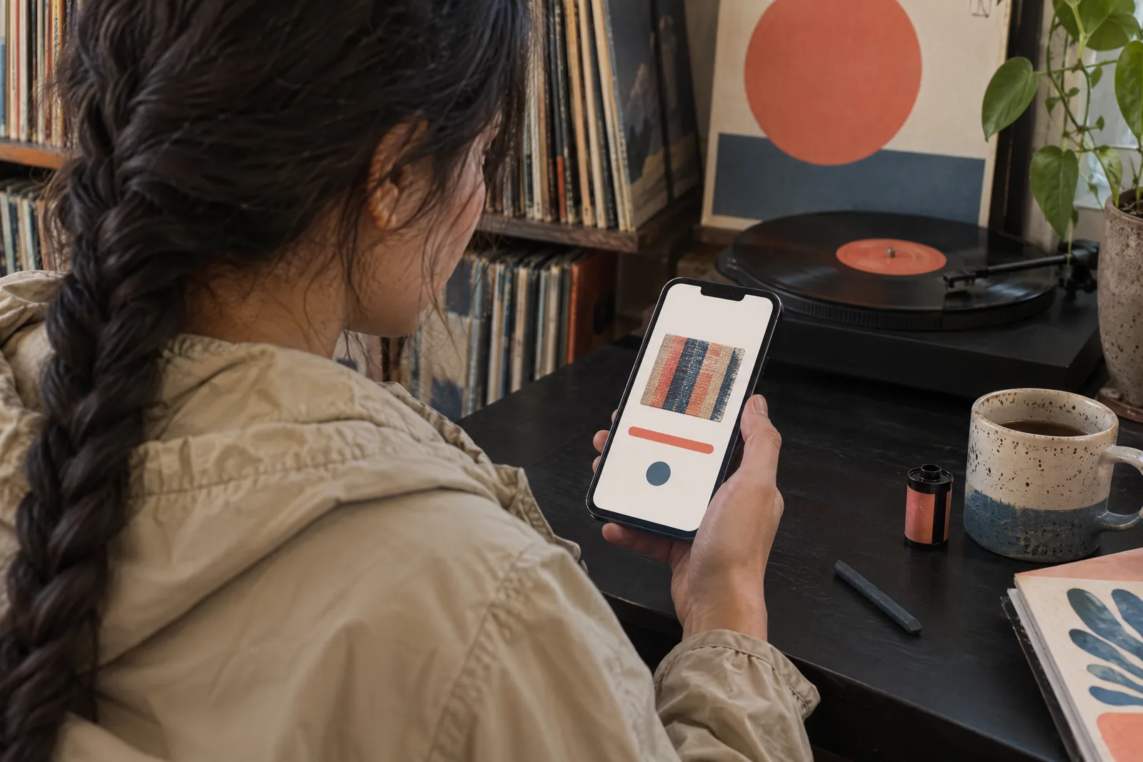 A woman in her twenties with a long dark braid in a sand rain jacket at a record nook looks at a screen showing a video product for sale, with a charcoal stick beside them.