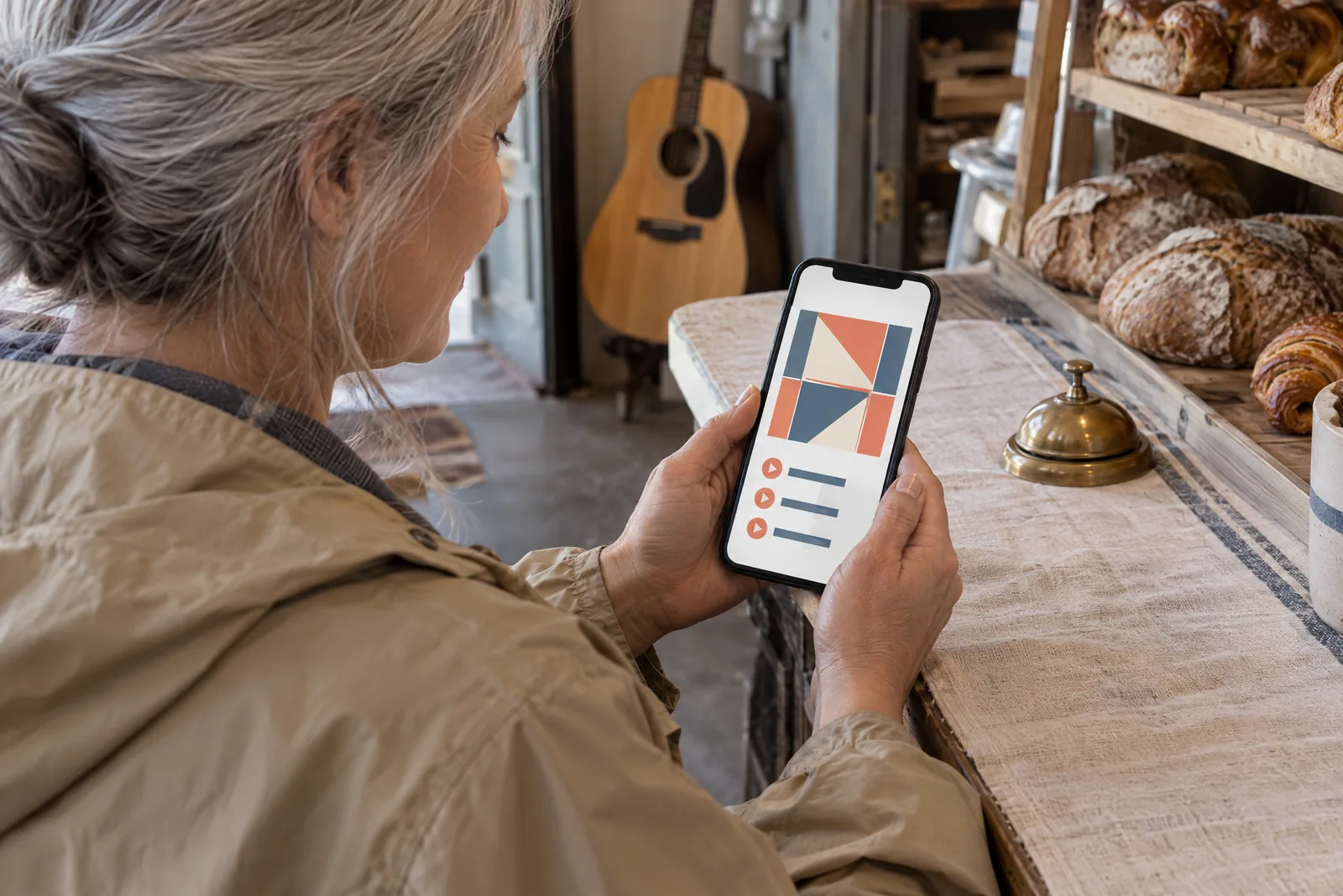 A woman in her fifties with silver hair in a low bun in a sand rain jacket at a bakery side counter looks at a screen showing a music product for sale, with a small brass bell beside them.