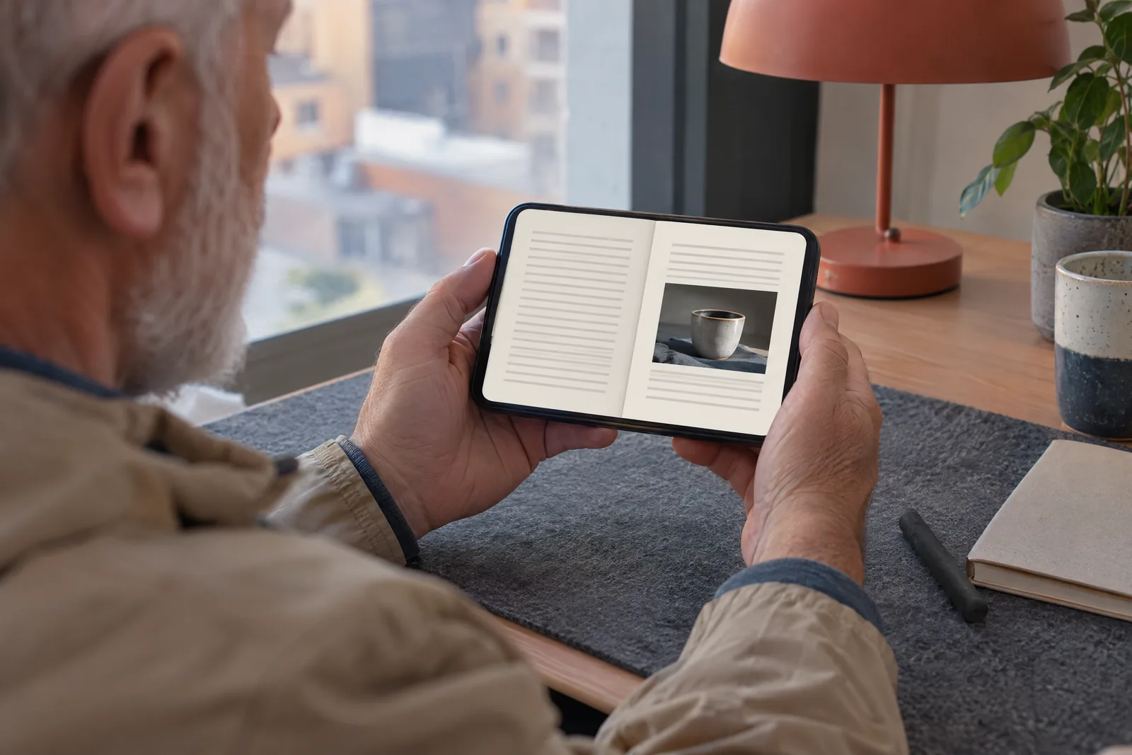 A man in his sixties with a close-cropped white beard in a sand rain jacket at a rooftop room looks at a screen showing a book product for sale, with a charcoal stick beside them.