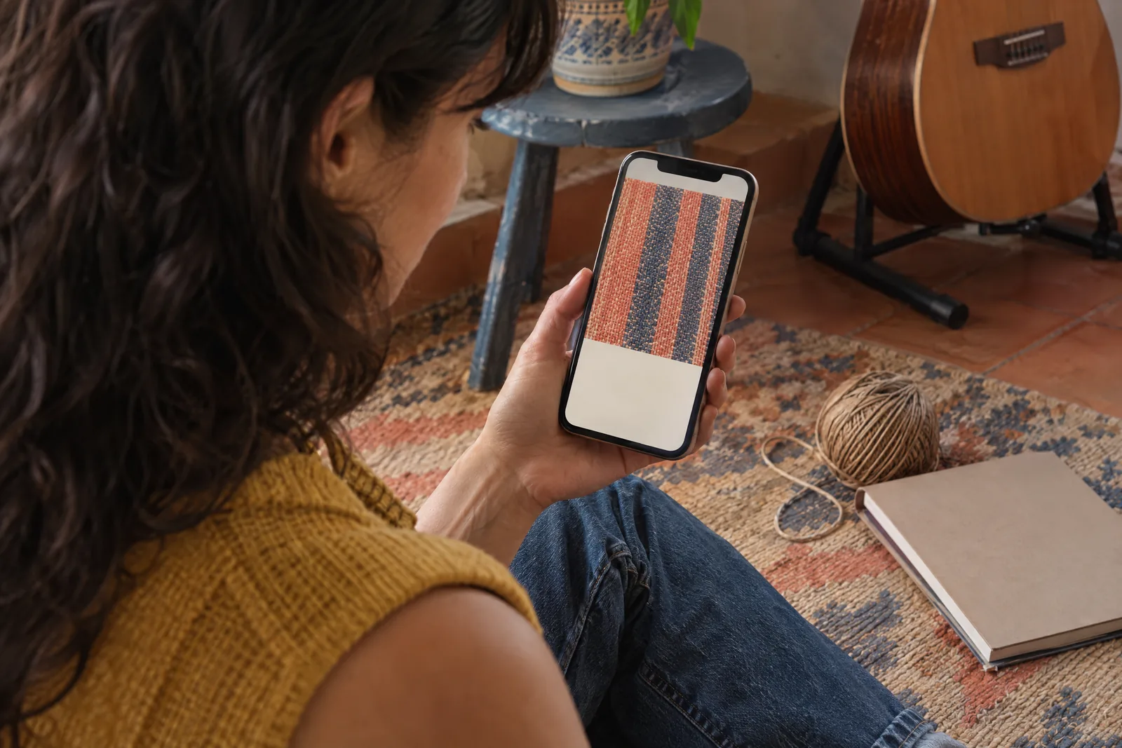 A woman in her thirties with dark wavy hair in a mustard knit vest at a rehearsal corner looks at a screen showing a ebook product for sale, with a ball of twine beside them.