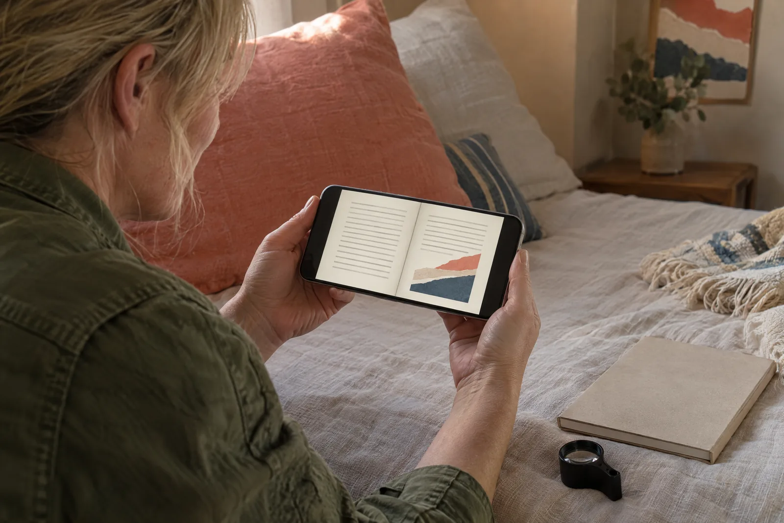 A woman in her forties with blonde hair tied back in a olive field jacket at a bedroom alcove looks at a screen showing a book product for sale, with a film loupe beside them.
