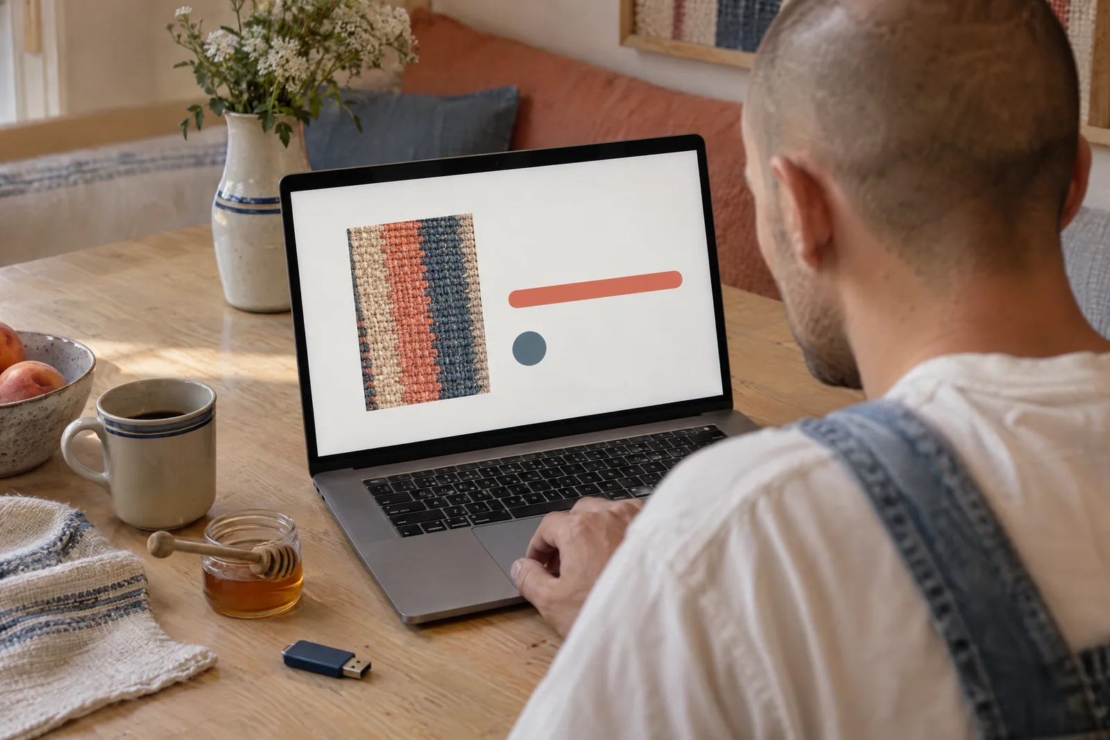 A man in his twenties with a shaved head in a denim overalls at a dining alcove looks at a screen showing a software product for sale, with a honey dipper beside them.