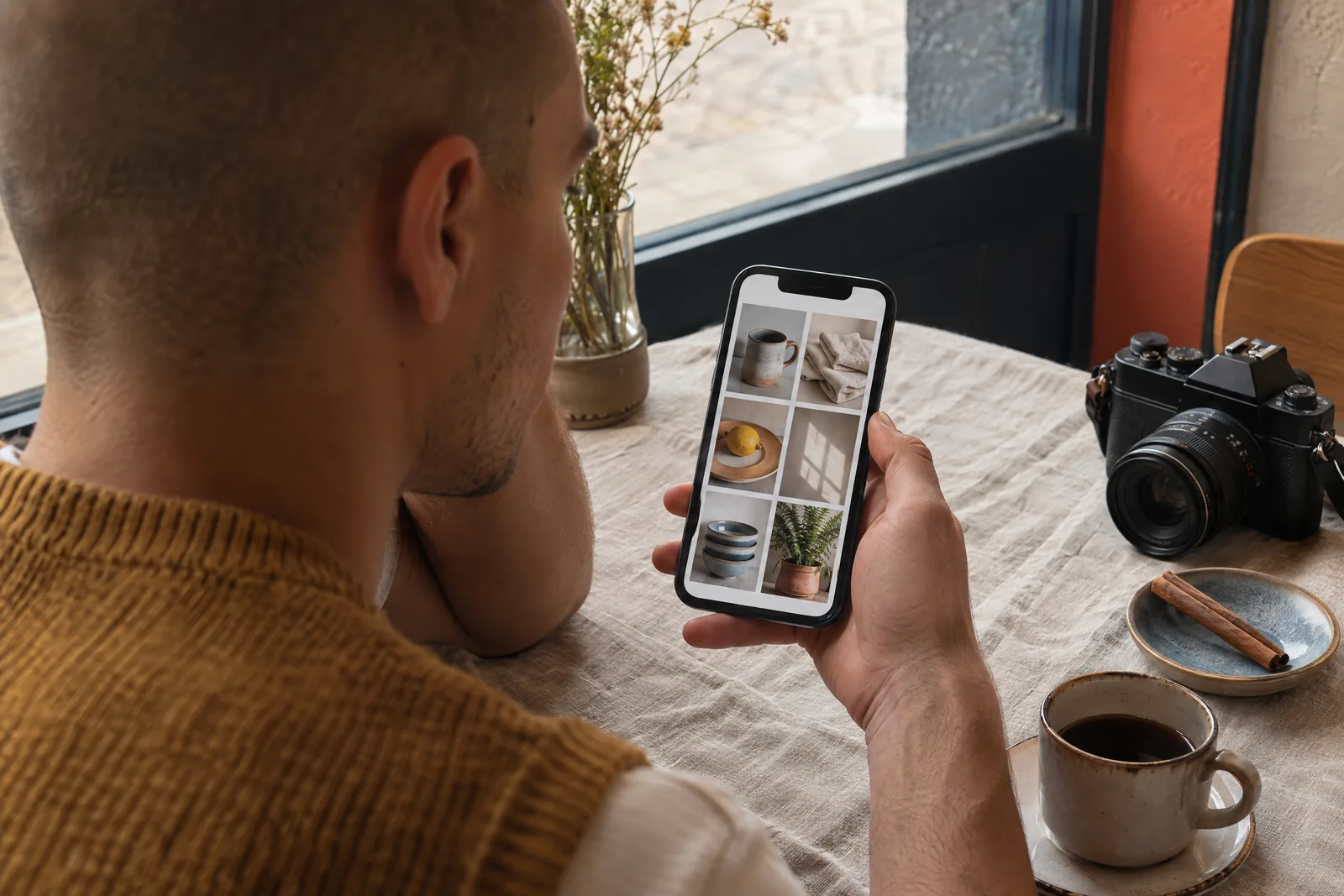 A man in his twenties with a shaved head in a mustard knit vest at a cafe window looks at a screen showing a photo product for sale, with a cinnamon stick beside them.