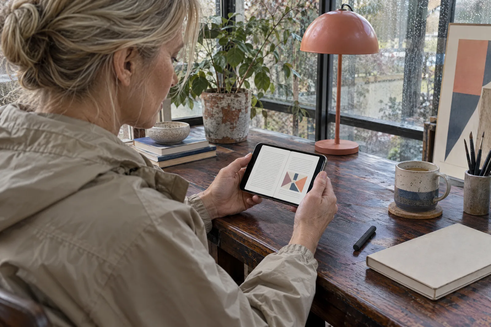 A woman in her forties with blonde hair tied back in a sand rain jacket at a glass studio looks at a screen showing a book product for sale, with a charcoal stick beside them.