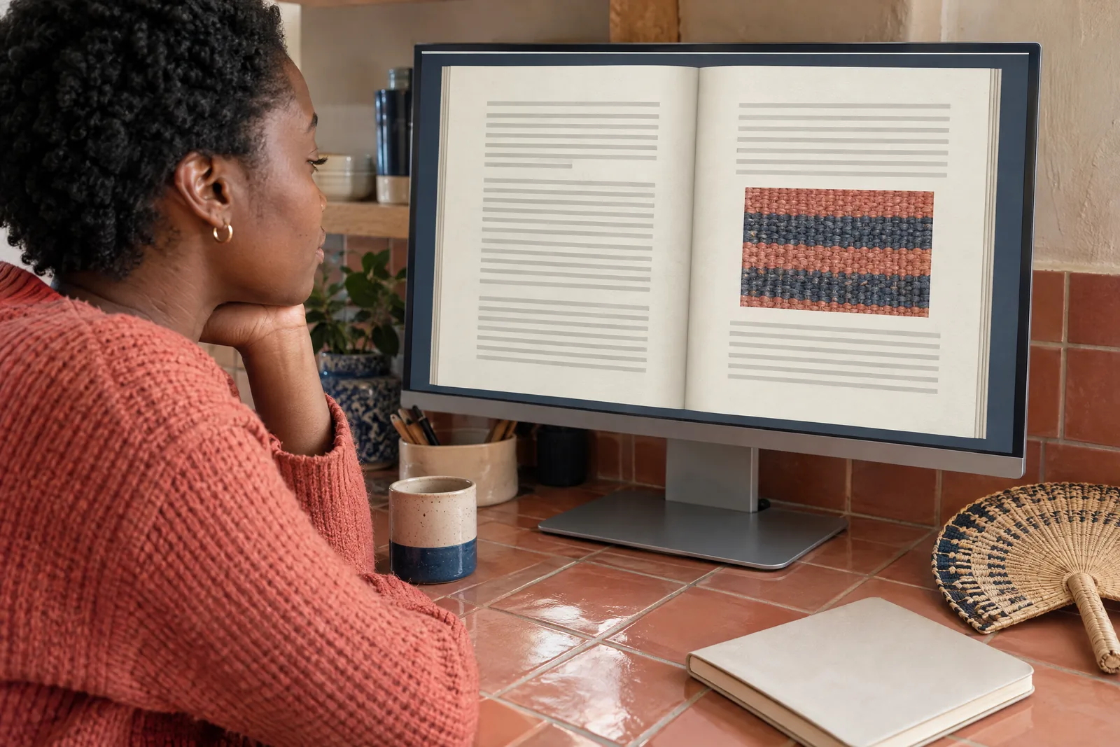 A black woman in her twenties with short natural hair in a coral knit sweater at a kitchen nook looks at a screen showing a book product for sale, with a paper fan beside them.