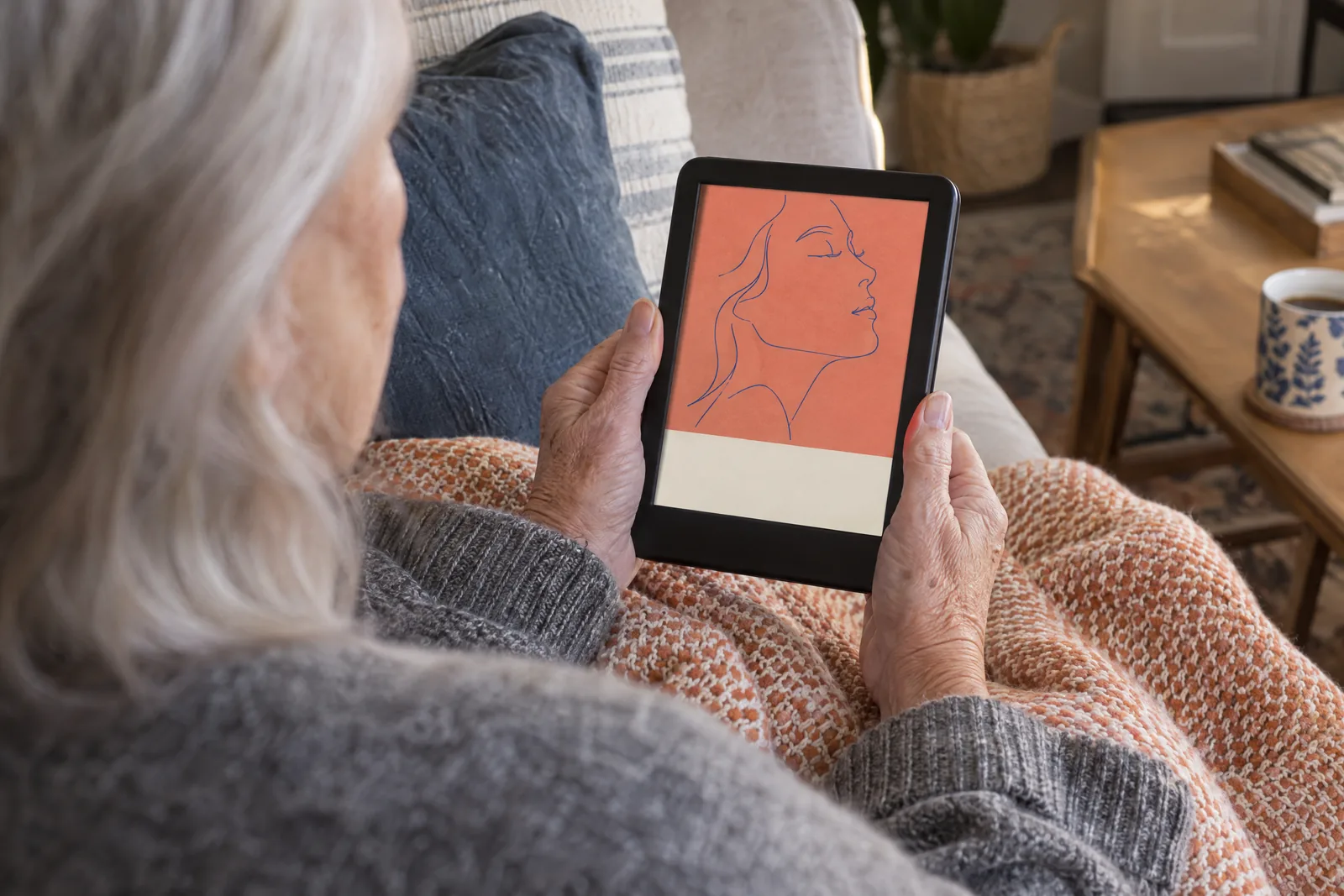 Over-the-shoulder view of an older woman on a sofa holding an e-reader that shows an ebook cover with a line portrait.