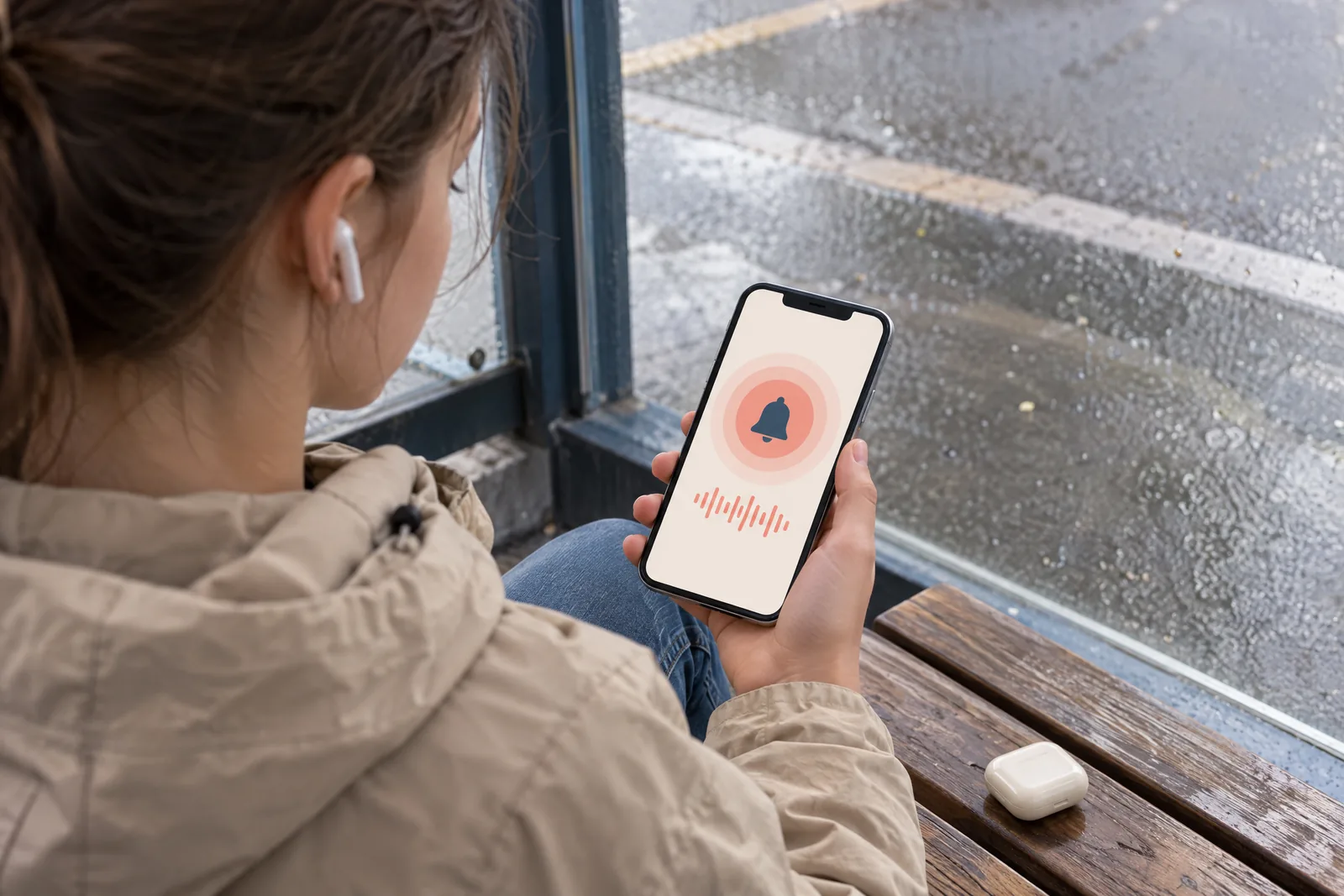 Over-the-shoulder view of a teenager on a bus stop bench holding a phone that shows a ringing screen with coral rings.