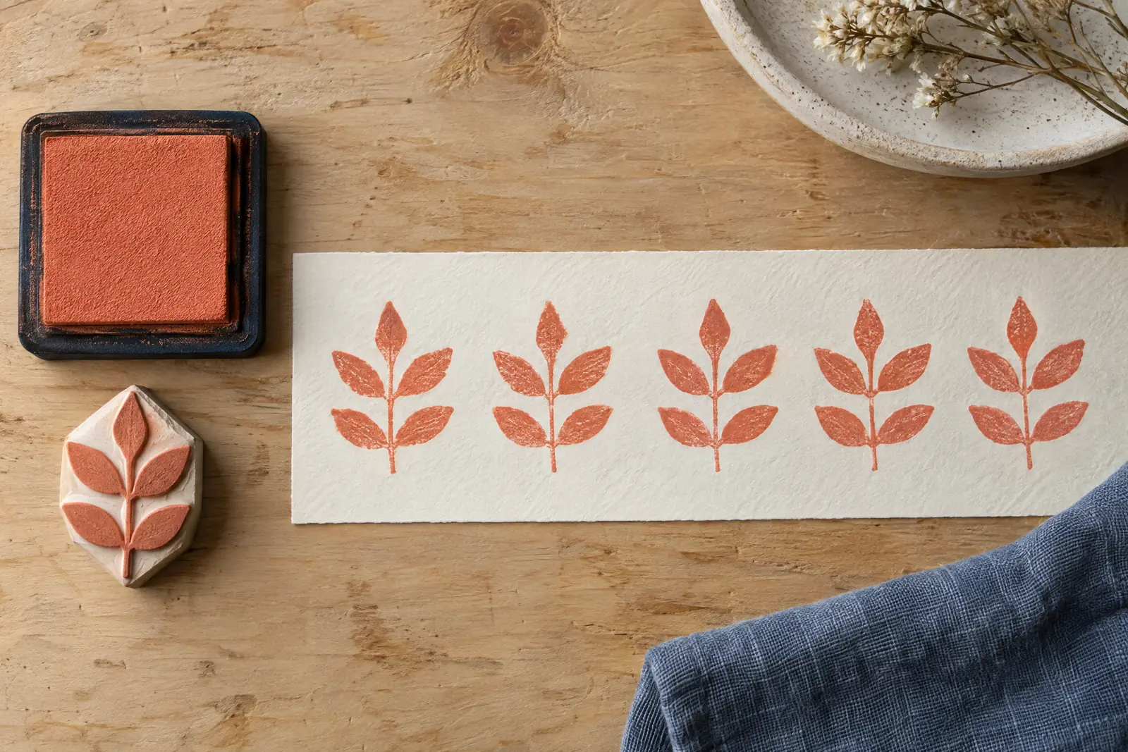 Overhead still life of one carved stamp and a coral ink pad beside a paper strip printed with a row of identical coral leaves.